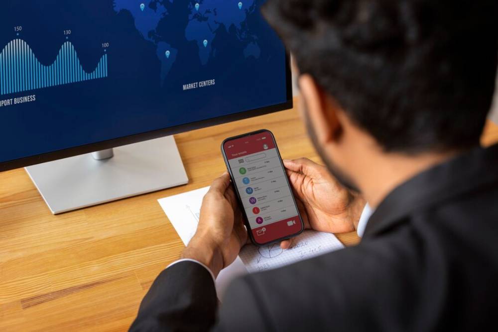 A man in a suit uses a smartphone while analyzing data on a computer screen displaying a world map and statistics.