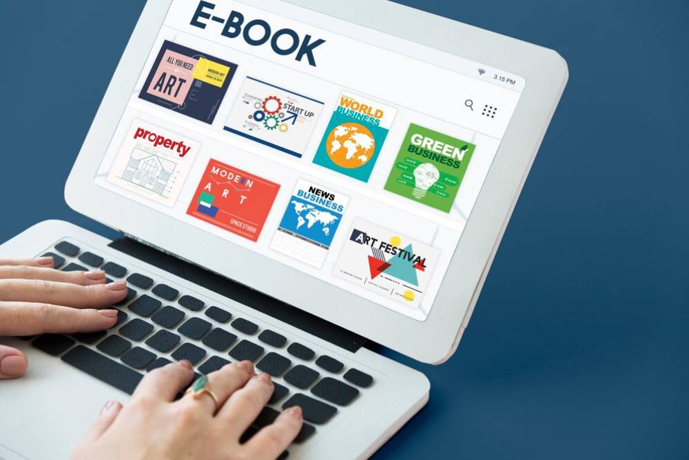 A person’s hands on a laptop keyboard displaying a digital bookshelf of colorful e-books on various topics.