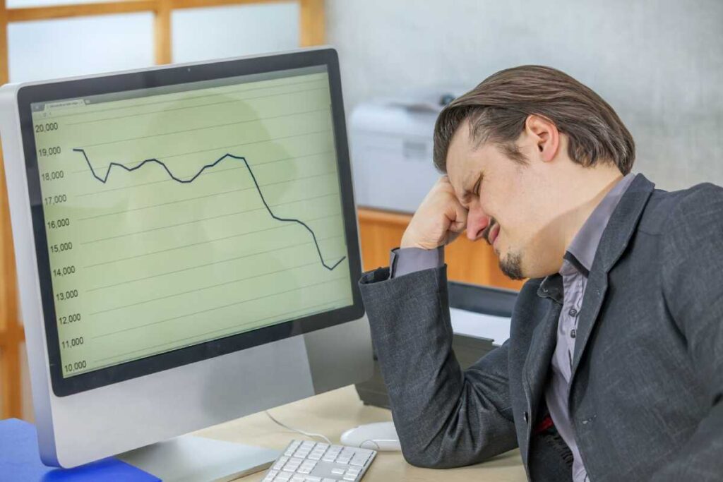 A person in a suit rests their head on their hand, looking at a computer screen displaying a declining graph and financial data.