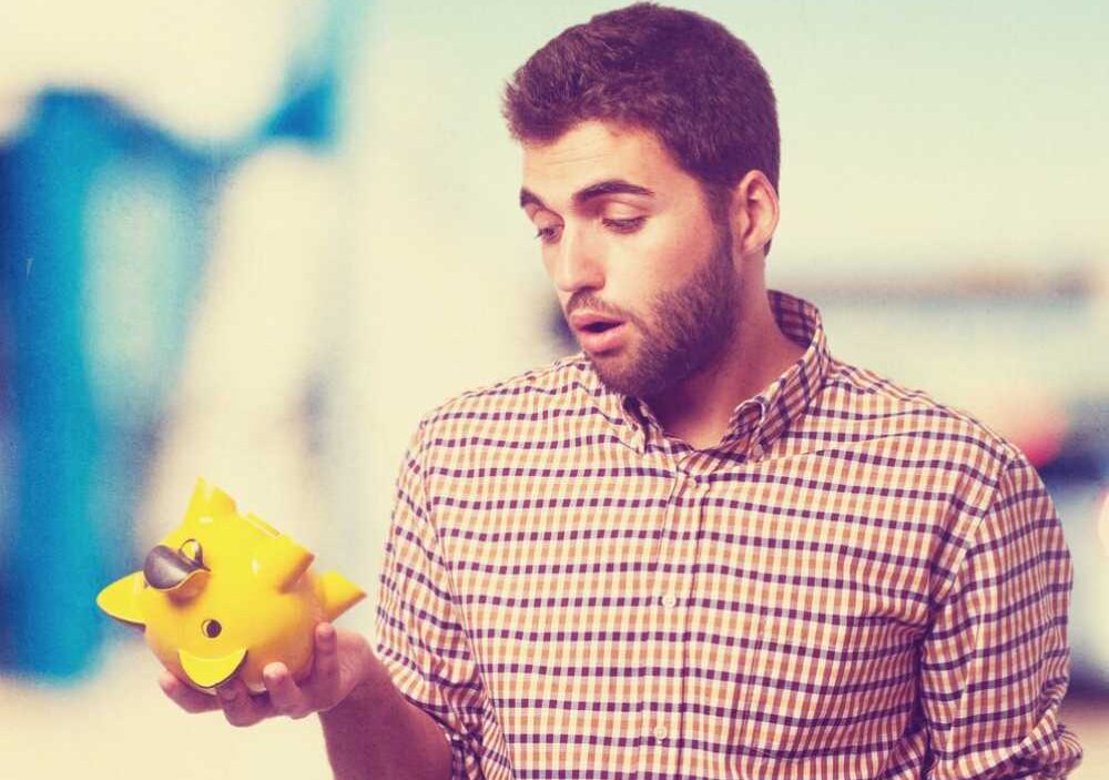 A man in a checked shirt holds a yellow piggy bank, appearing to ponder financial decisions against a blurred background.
