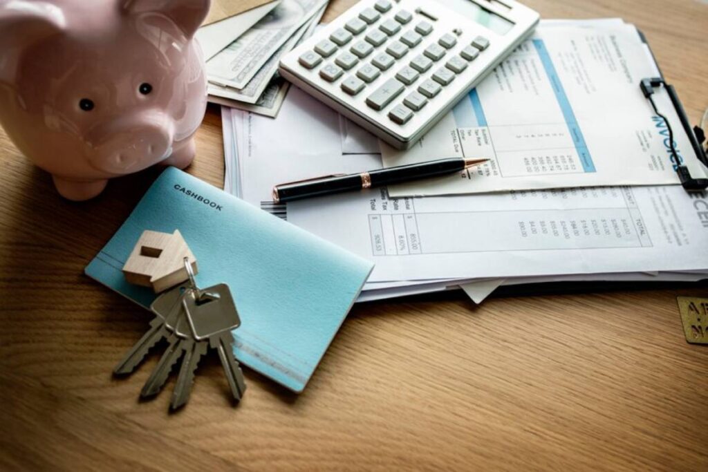 A pink piggy bank, keys, a cashbook, bills, a calculator, and a pen arranged on a wooden desk, symbolizing estate planning and personal finance management.