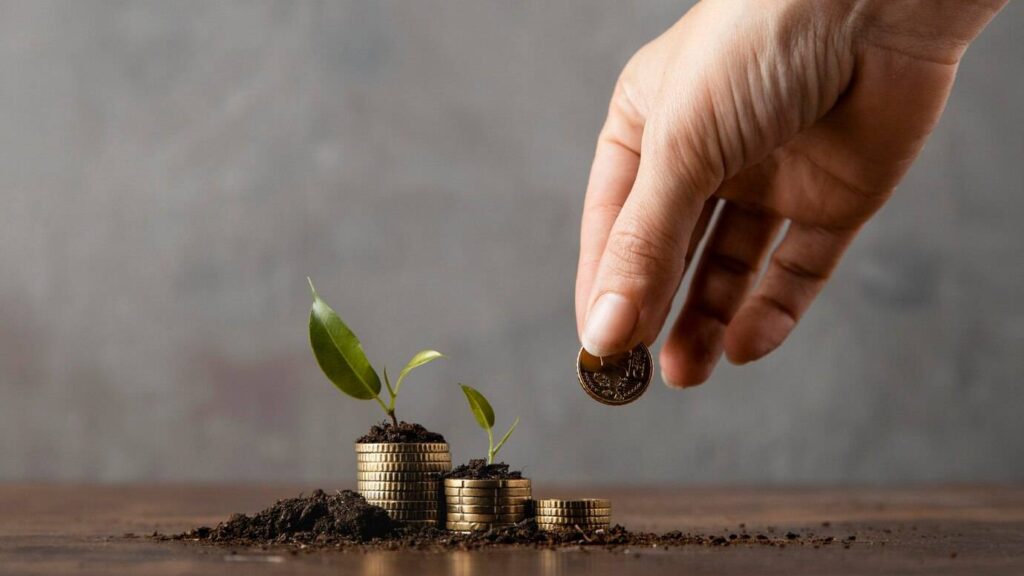 A hand dropping a coin onto soil with growing plants and stacks of gold coins, symbolizing investment and growth.