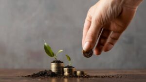 A hand dropping a coin onto soil with growing plants and stacks of gold coins, symbolizing investment and growth.