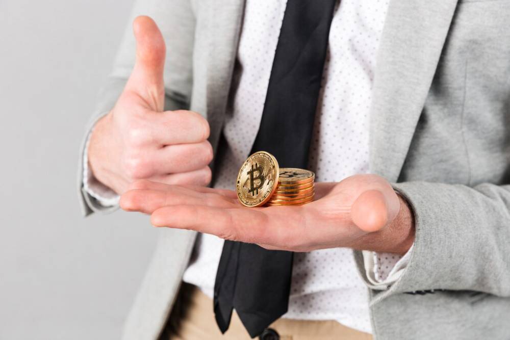 A businessman in a gray suit gives a thumbs up while holding a stack of Bitcoin coins in one hand and a Bitcoin in the other.
