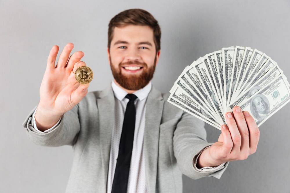 A person in a suit holds a Bitcoin in one hand and a fan of hundred-dollar bills in the other, against a gray background.