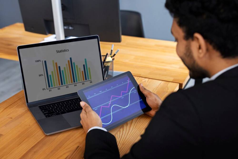 A person in a suit analyzes data on a tablet, while a laptop displays colorful statistical graphs on a wooden desk.