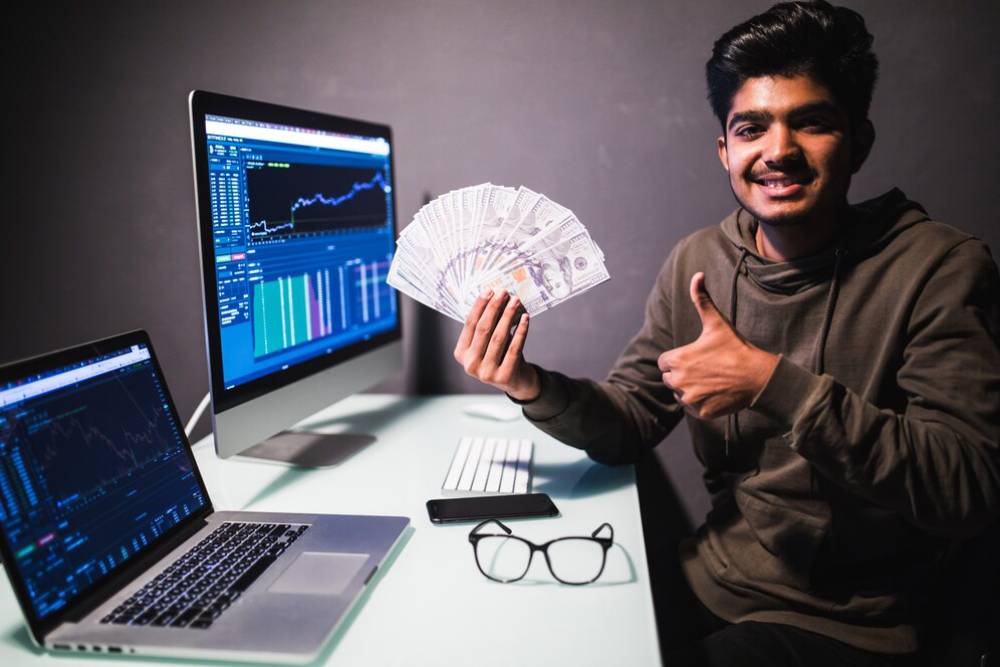 A person holds a fan of cash and gives a thumbs-up, sitting at a desk with two laptops displaying stock charts and data.