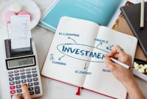 A hand writing "INVESTMENT" in a notebook, surrounded by keywords like "ACCOUNTING," "FINANCE," and "FUNDING," with a calculator nearby.