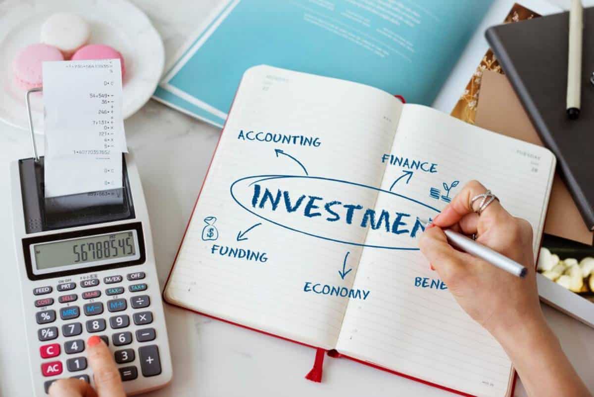 A hand writing "INVESTMENT" in a notebook, surrounded by keywords like "ACCOUNTING," "FINANCE," and "FUNDING," with a calculator nearby.
