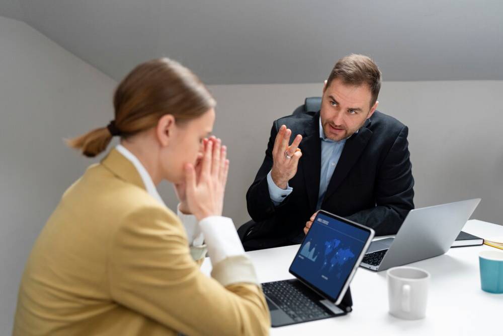 A professional meeting in a modern office setting, featuring two individuals engaged in discussion with laptops and a notepad on the table.