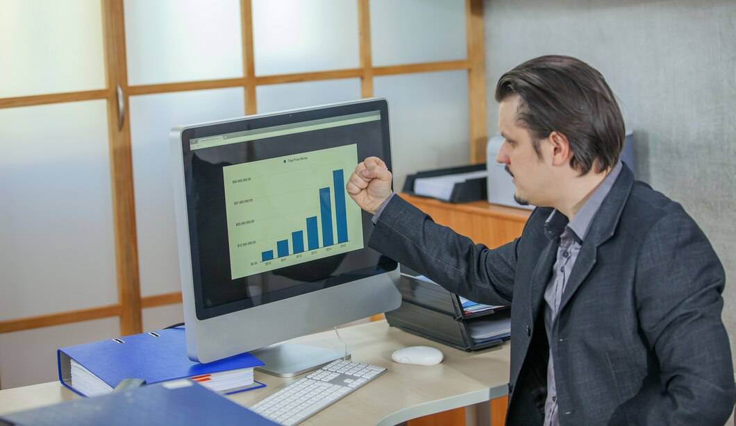 A man in a suit enthusiastically points to a rising bar graph on a computer screen, indicating success in data analysis.