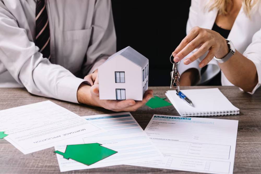 A man holds a small model house while a woman hands him keys, surrounded by mortgage documents and a notebook on a wooden table.