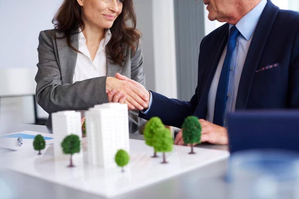 A business meeting featuring two professionals shaking hands over a model cityscape with miniature buildings and trees on the table.