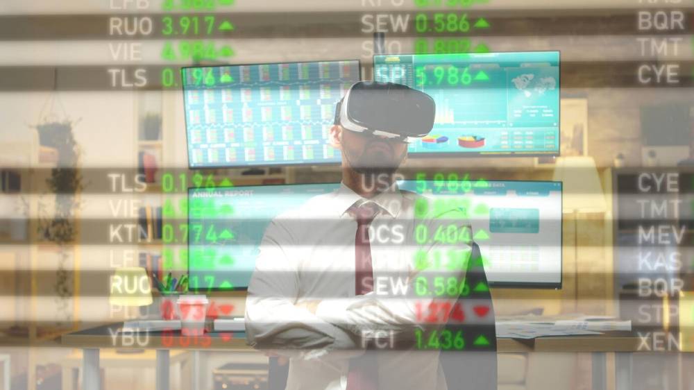 A businessman wearing a VR headset stands in front of multiple monitors displaying stock market data and reports.
