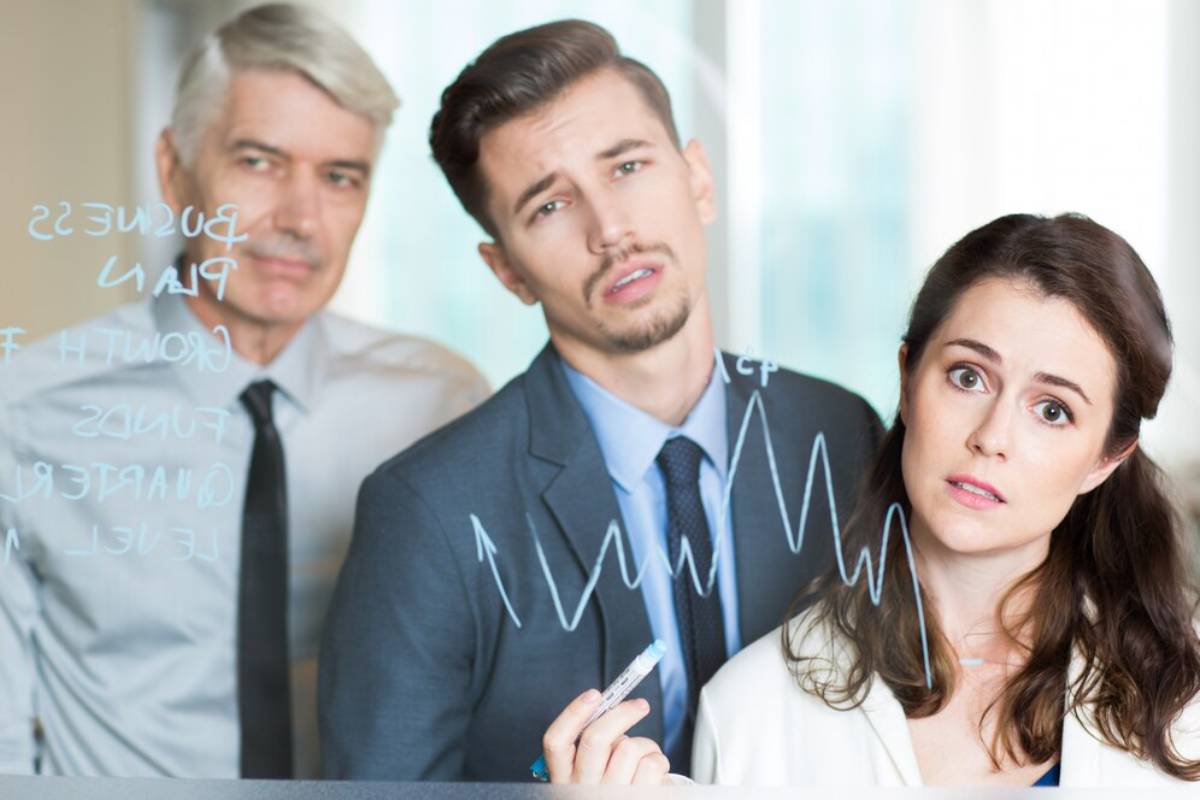 A group of professionals discusses data on a glass board, marked with charts and notes, in a bright office setting.