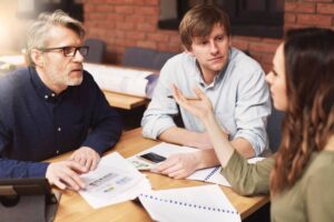 A group of three people engaged in a discussion around a table, surrounded by papers and designs, with a warm brick background.