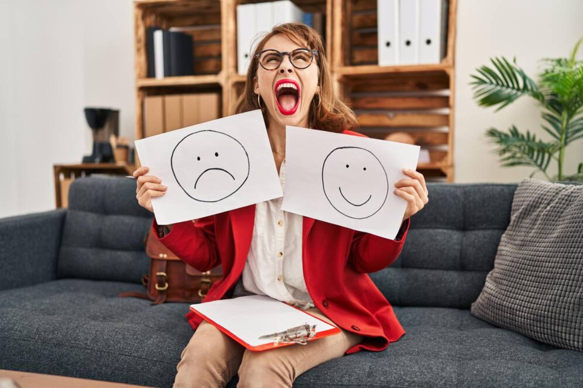 A person in a red blazer holds two drawings: one with a sad face and another with a happy face, sitting on a gray couch.