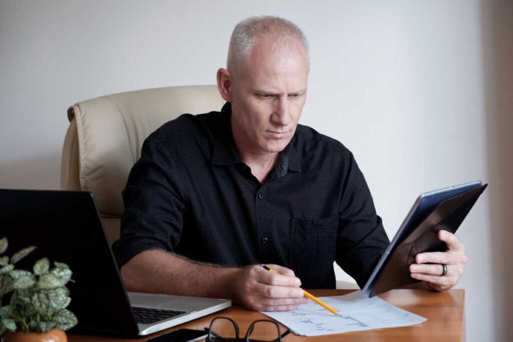 A man in a dark shirt sits at a desk with a laptop, tablet, and papers. A small plant is nearby.