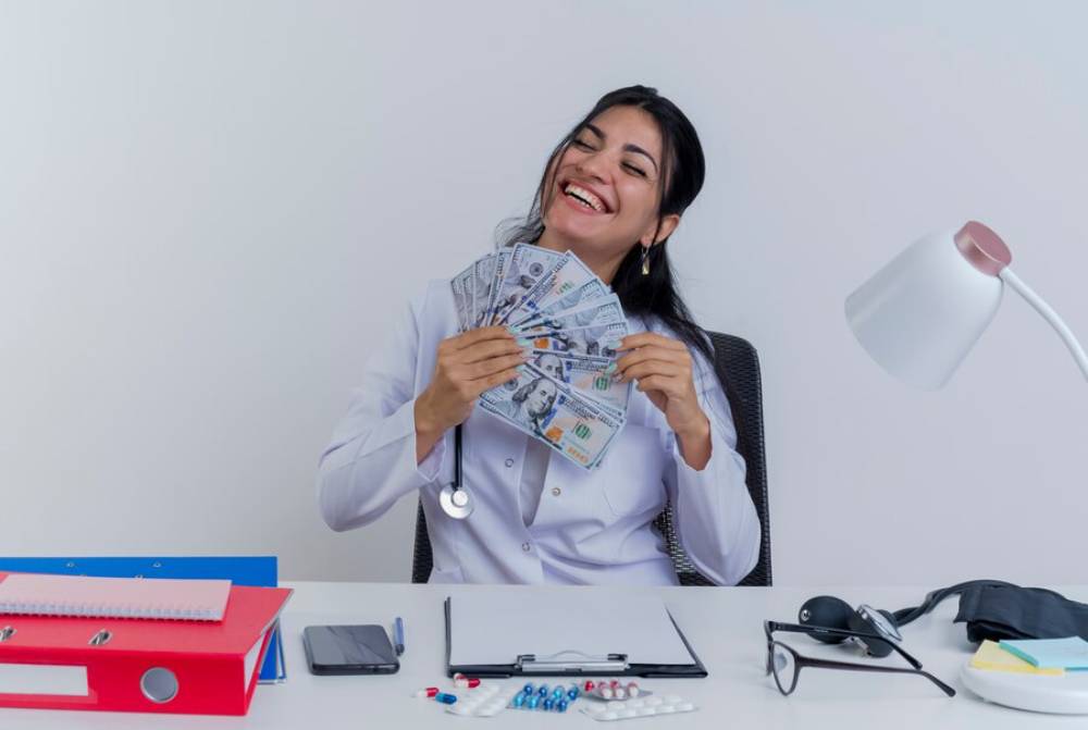A person in a white coat holding a fan of cash, with a desk cluttered with files, pens, and medical supplies.