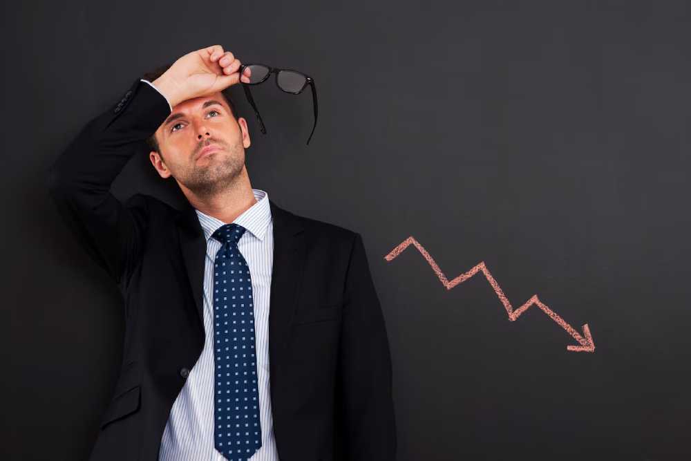 A businessman holds glasses above his head, next to a downward trend line drawn on a chalkboard symbolizing loss in investment.
