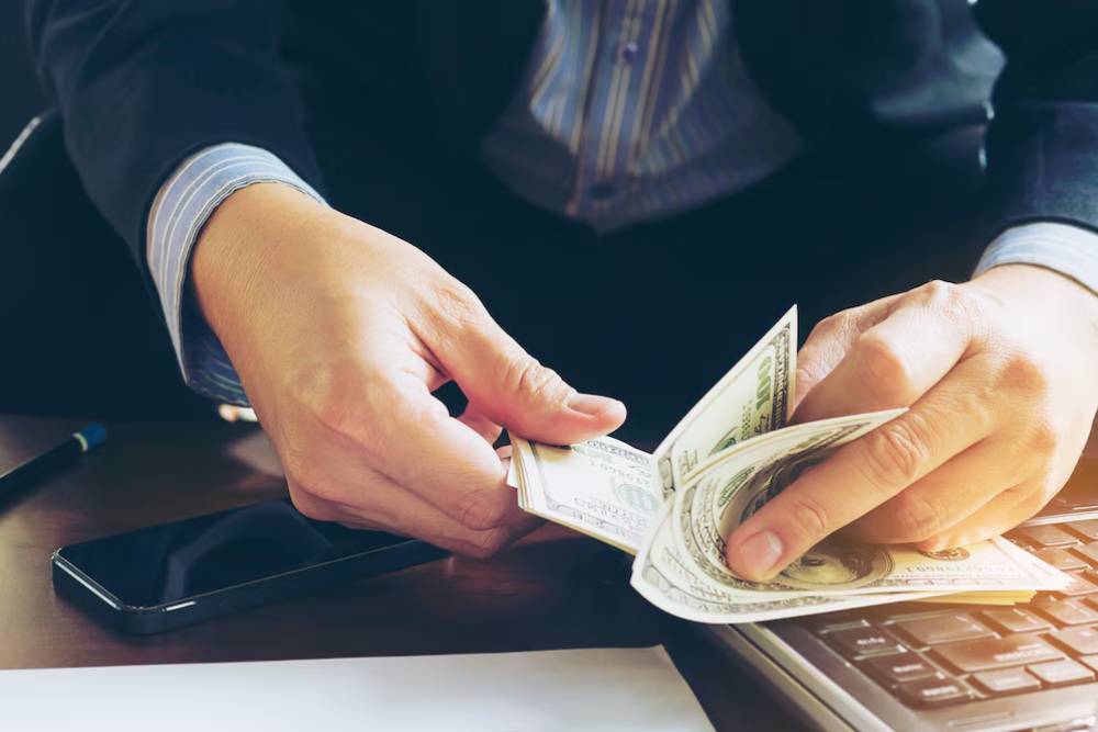 A businessman counts cash on a desk next to a laptop and smartphone, focusing on the bills in his hands.