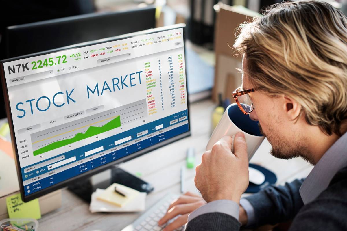 A person holding a coffee mug sits in front of a computer displaying stock market data and charts.