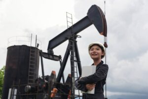 A worker in a hard hat stands with a clipboard in front of an oil rig, under a cloudy sky.