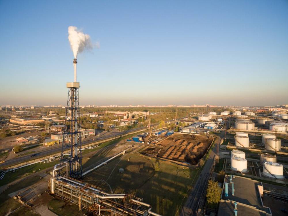 Aerial view of an industrial facility with smoke rising from a tower, surrounded by storage tanks and infrastructure against a clear sky.