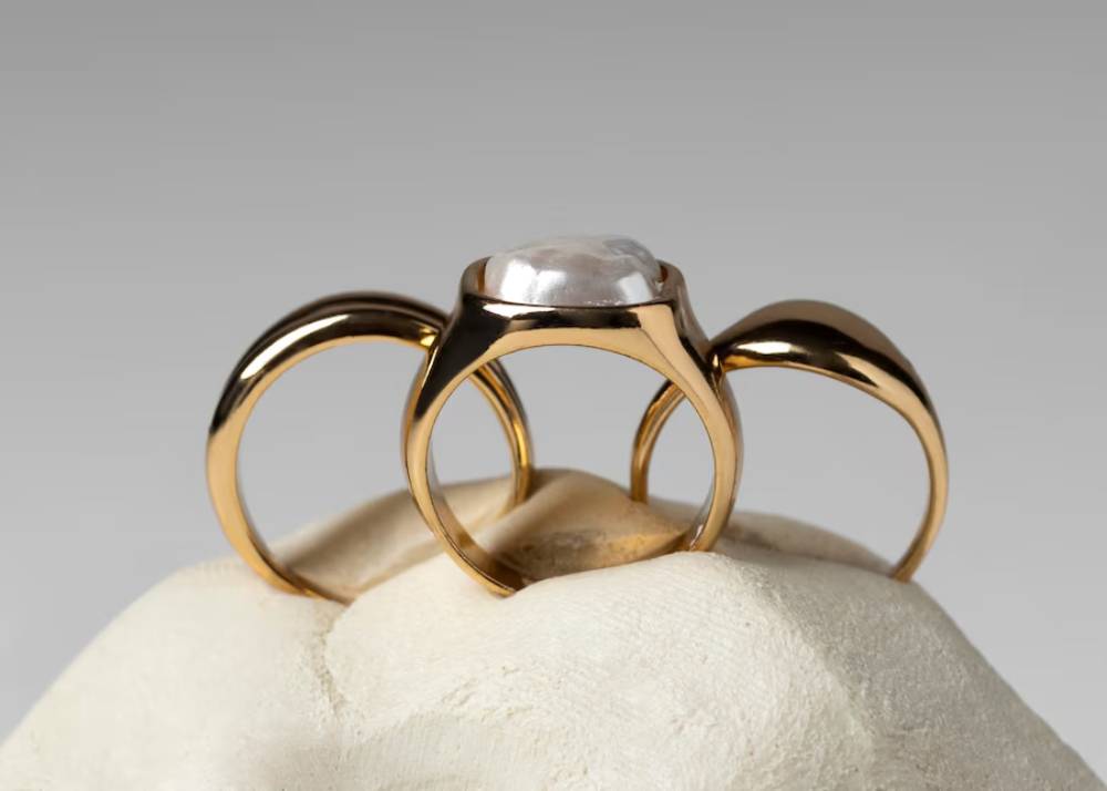 A trio of elegant gold rings, one adorned with a large, lustrous gemstone, displayed atop a smooth, pale stone.