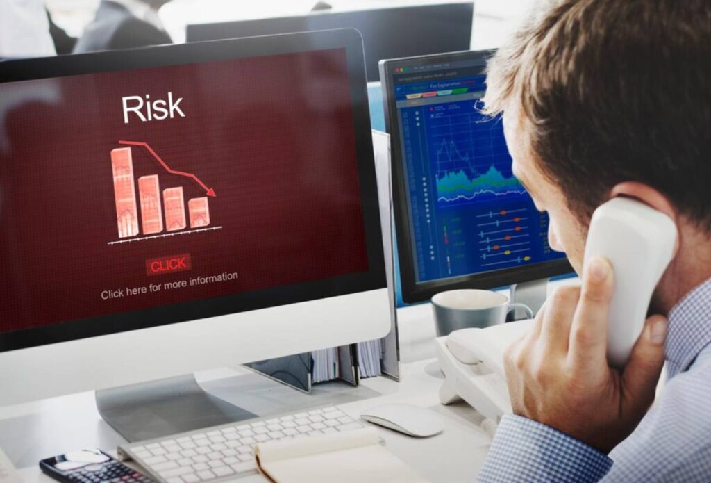 A businessman on a phone, analyzing a computer screen displaying a downward risk graph and market data charts.