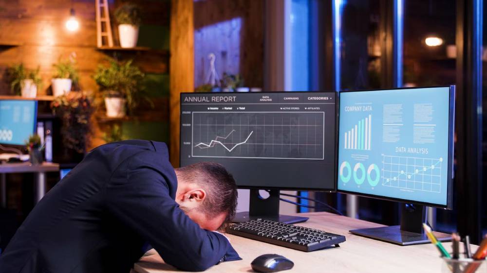 A frustrated businessman with his head down on a desk, surrounded by dual monitors displaying data analysis and reports.
