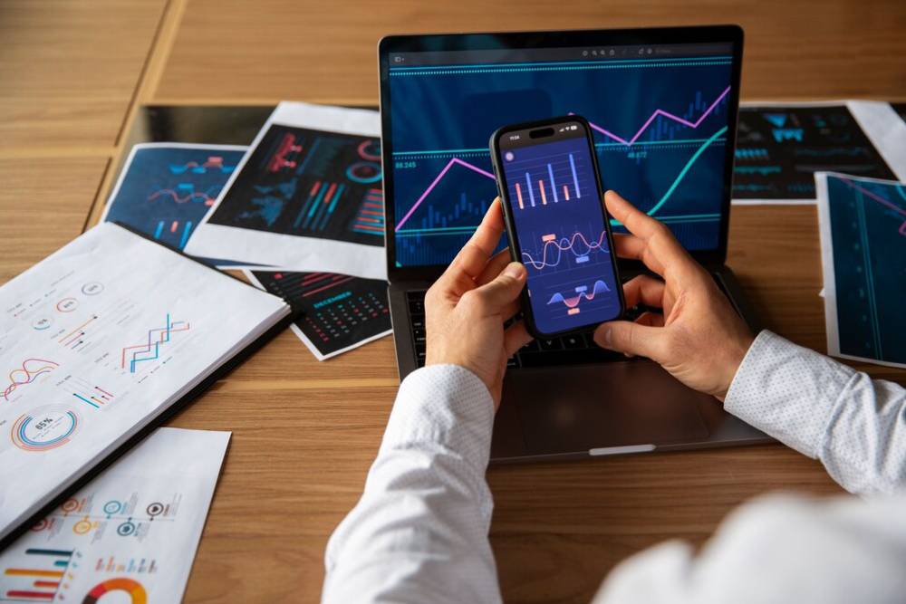 A person analyzing graphs and data on a smartphone while working on a laptop, with printed charts spread across a wooden desk.
