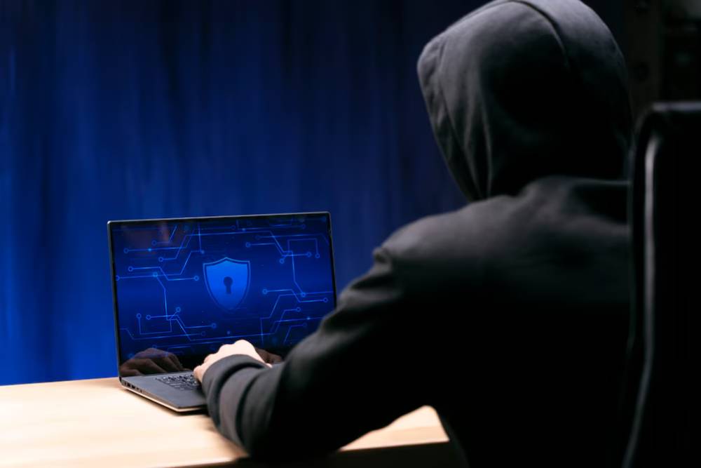 A hooded figure sits at a desk, focused on a laptop displaying a shield icon, symbolizing cyber security or hacking.