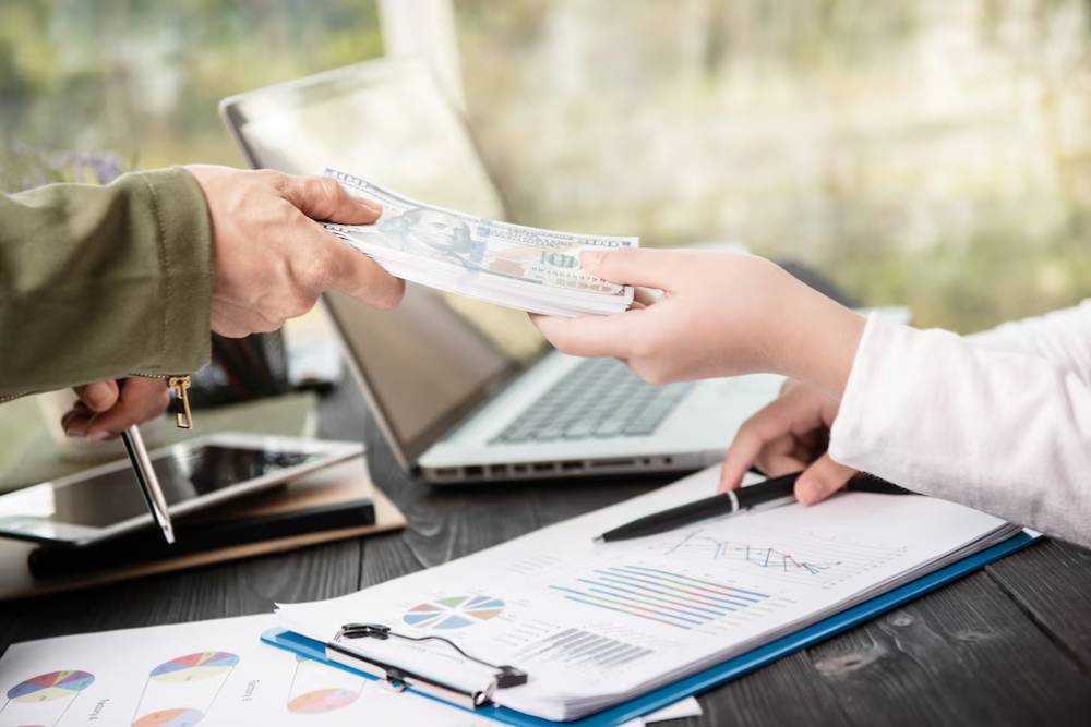 A person hands cash to another while discussing financial documents and charts on a desk with a laptop and tablet nearby.