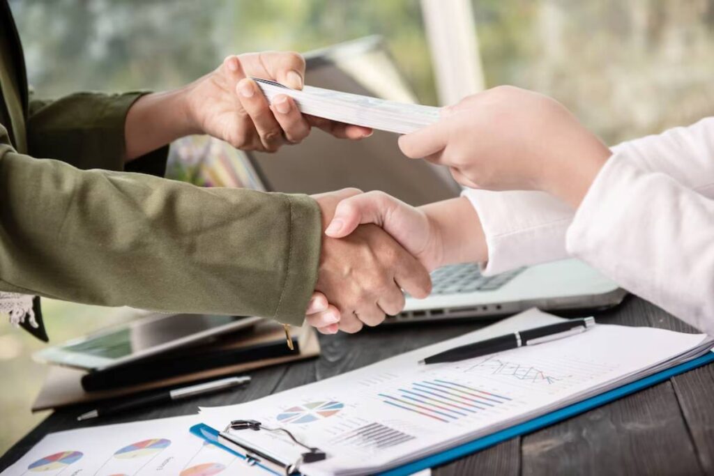 Two individuals shake hands while exchanging money at a desk covered with business reports and a laptop in the background.
