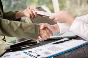 Two individuals shake hands while exchanging money at a desk covered with business reports and a laptop in the background.