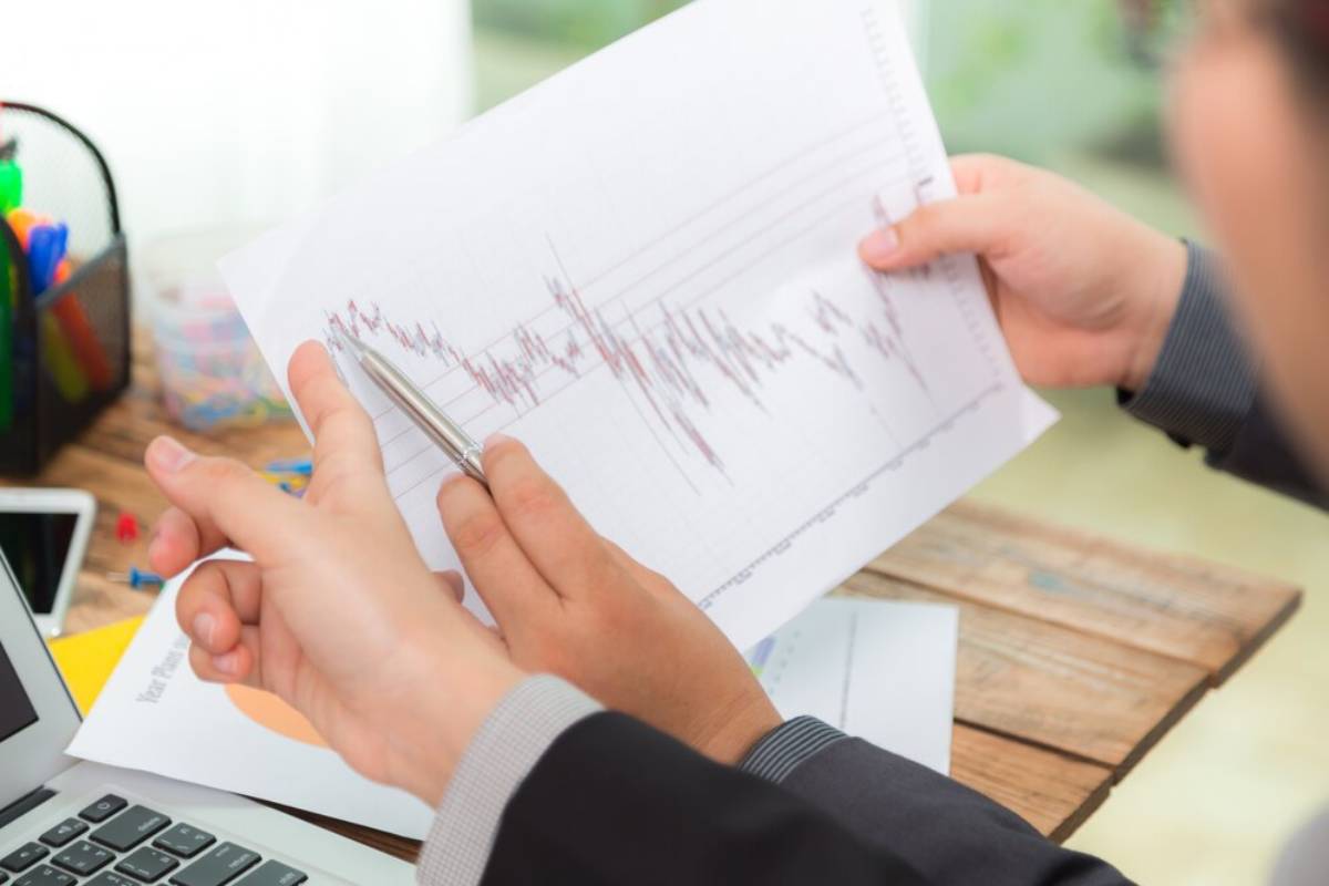 A person in a suit holds a financial graph with a pen, analyzing data on a wooden desk with office supplies and a laptop in the background.
