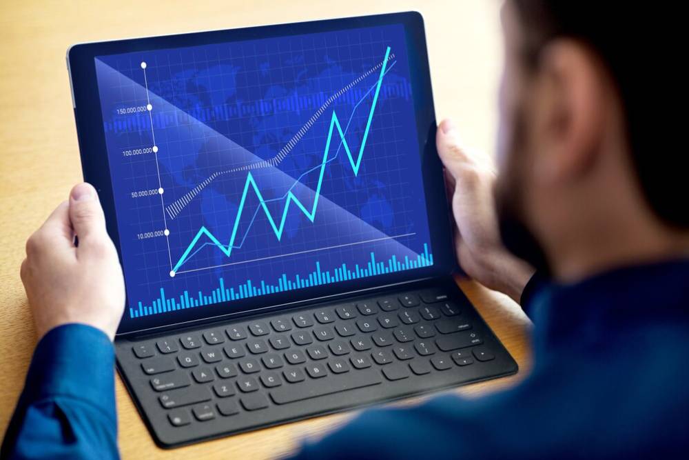A person viewing a tablet displaying a blue graph with rising trends and data bars on a wooden table, indicating financial analysis.