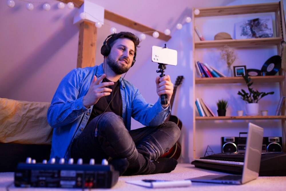 A person in headphones sits on the floor, holding a smartphone on a stick, surrounded by musical instruments and a laptop.