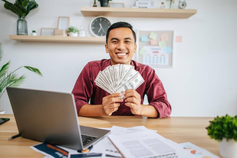 A person in a maroon shirt holds a fan of cash in front of a laptop on a desk, surrounded by documents and a potted plant.