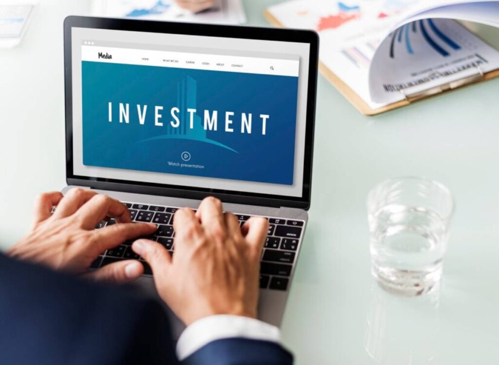 A person types on a laptop displaying an "Investment" webpage, with a glass of water nearby.