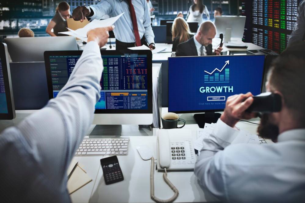 A busy trading room with multiple computer screens displaying financial data, while employees engage in discussions and phone calls.