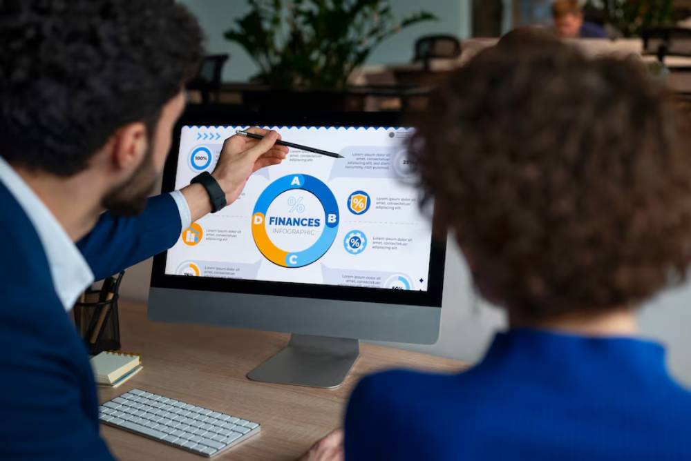 A person points at a finance infographic on a computer screen, while another individual observes attentively in a modern office setting.