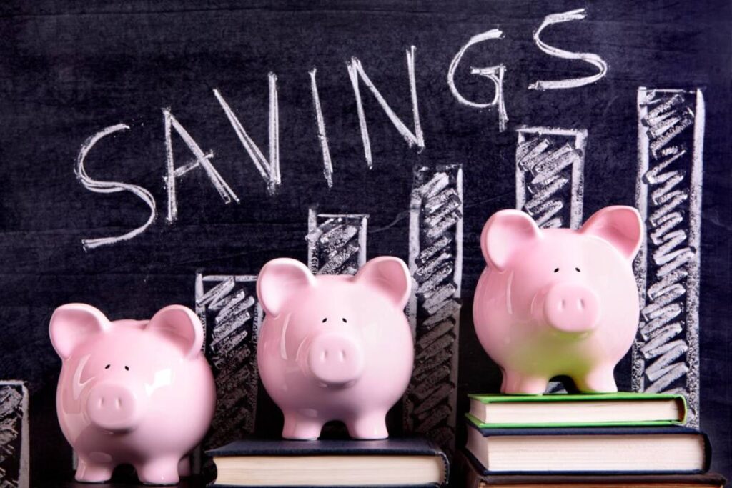 Three pink piggy banks are stacked on books, with a chalkboard diagram labeled "SAVINGS" illustrating financial growth.