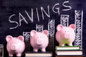 Three pink piggy banks are stacked on books, with a chalkboard diagram labeled "SAVINGS" illustrating financial growth.