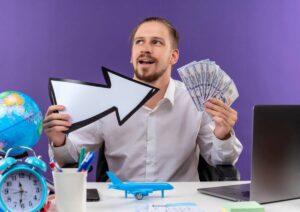 A person holds a large arrow sign and a fan of cash, surrounded by a globe, clock, and a small airplane on a desk.