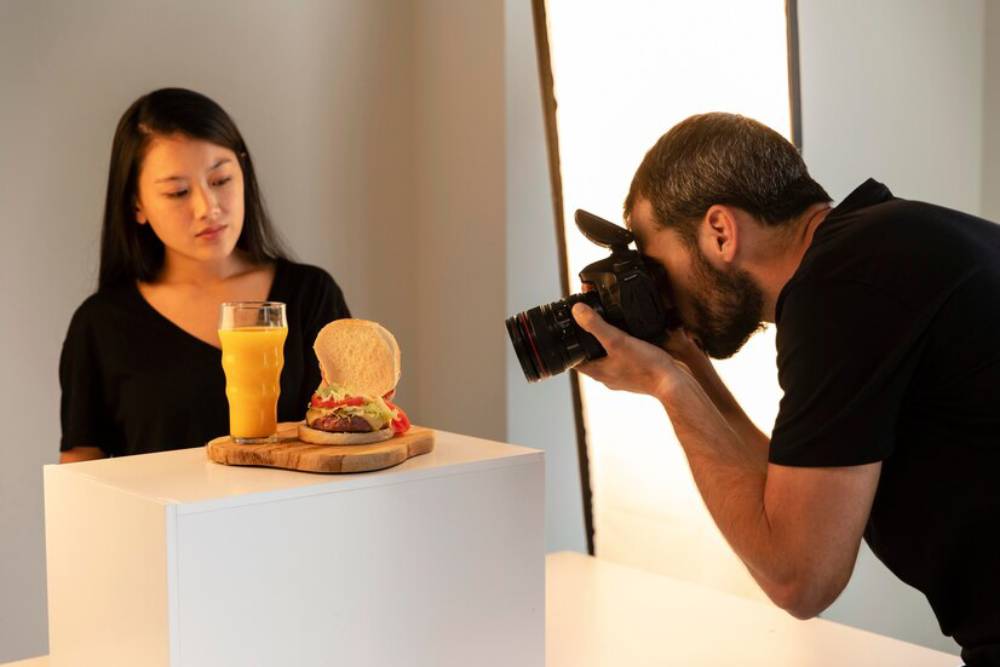 A photographer captures an appetizing burger and a glass of orange juice on a wooden board in a studio setting.