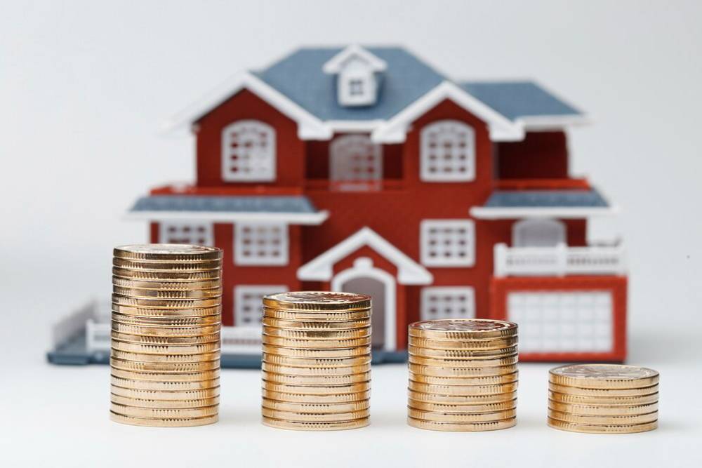 Stacks of gold coins increasing in height in front of a small red and white model house, symbolizing real estate investment.