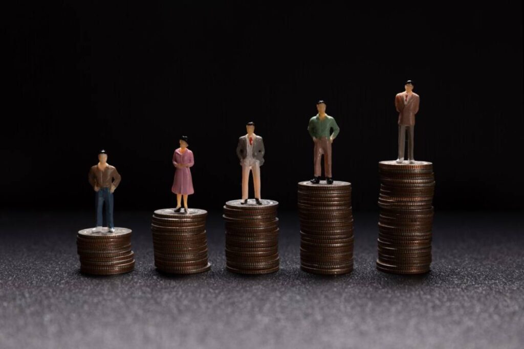 Five figurines stand atop stacks of coins, symbolizing wealth inequality and economic disparity against a dark background.