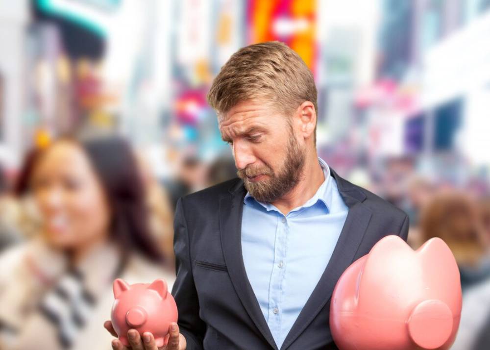 A man in a suit holds two pink piggy banks, standing in a busy, vibrant outdoor setting with blurred pedestrian activity around him.